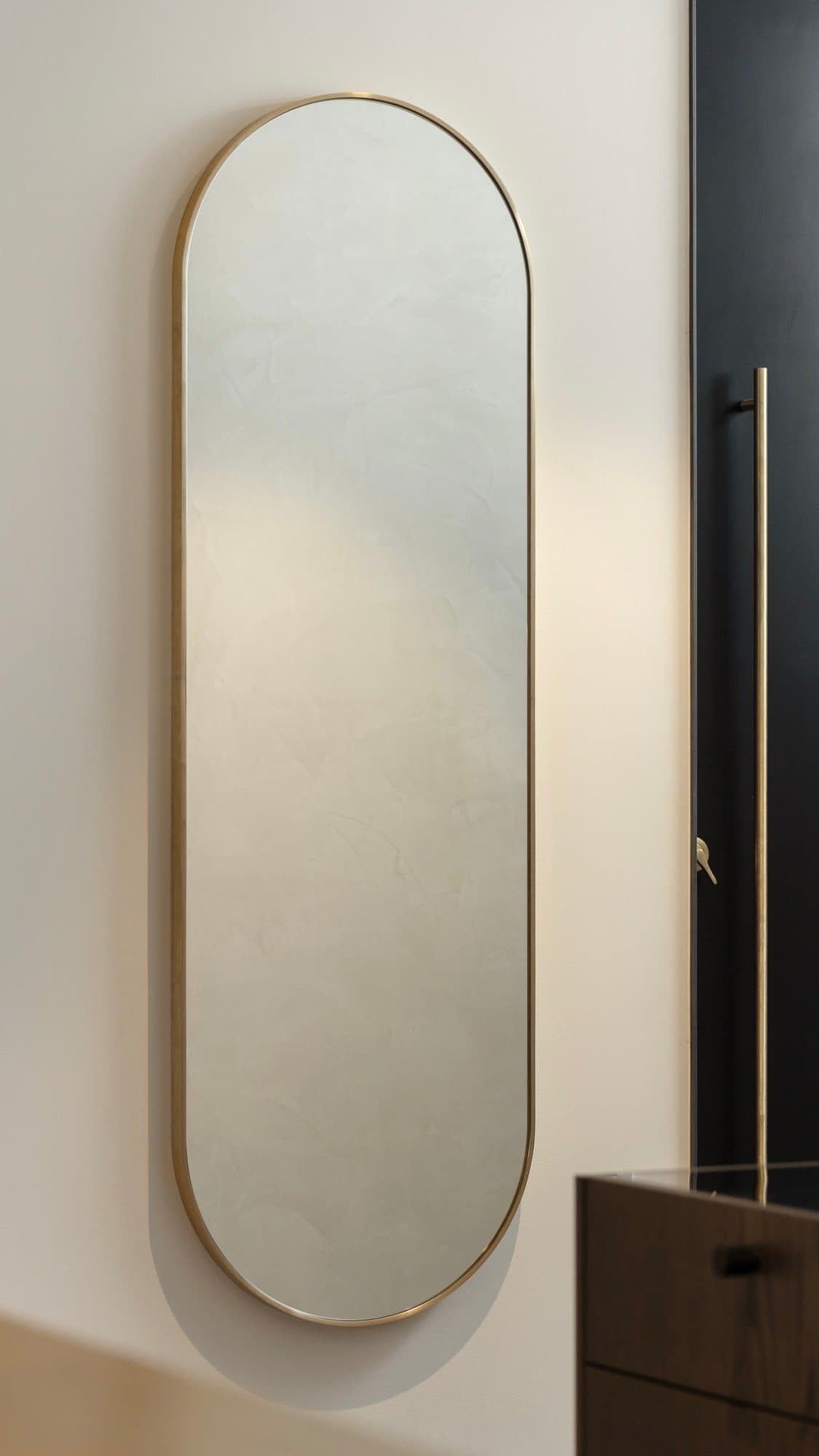 Tall brass pill mirror on a plaster wall