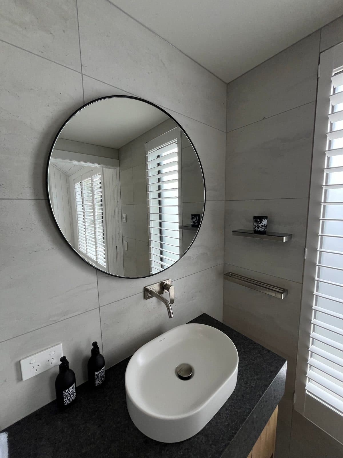 Round black mirror over an oval basin beside shutters